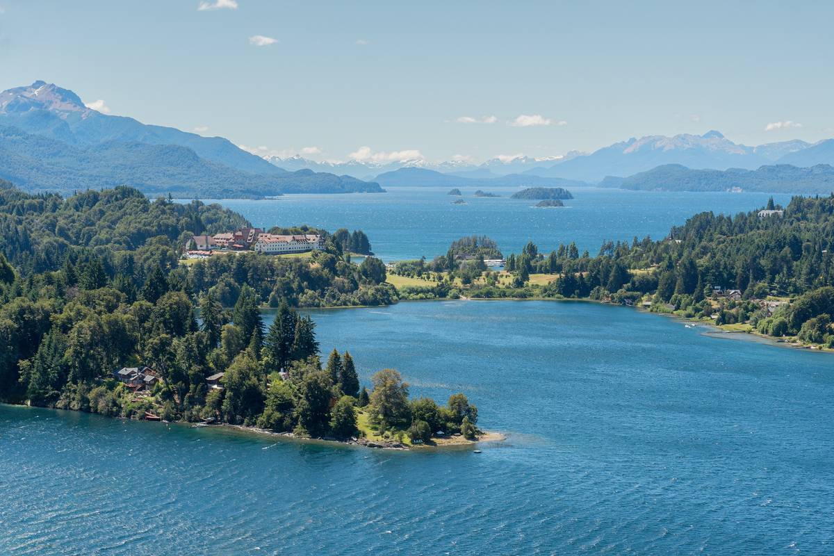Skogklädda halvöar och öar i en klarblå sjö vid Llao Llao, med Hotel Llao Llao bland träden och berg i diset bakom