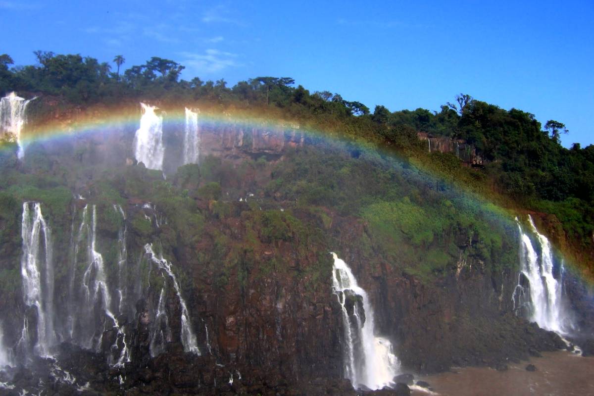 En regnbåge välver sig över flera vita vattenfall som störtar ner för en grön klippvägg i Iguazú under en klarblå himmel