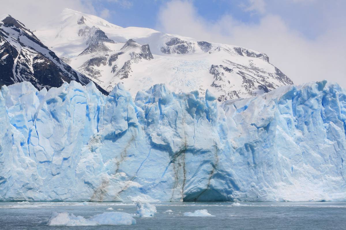 Perito Moreno-glaciärens isfront i vitt och isblått reser sig ur sjön framför ett snötäckt berg under en blå himmel