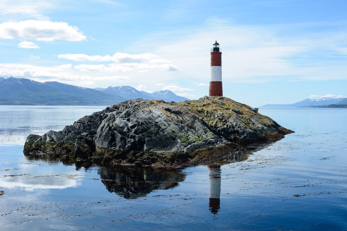 Den rödvita fyren Les Éclaireurs på en klippö i Beaglekanalen speglar sig i det stilla vattnet, med berg och vita moln i bakgrunden