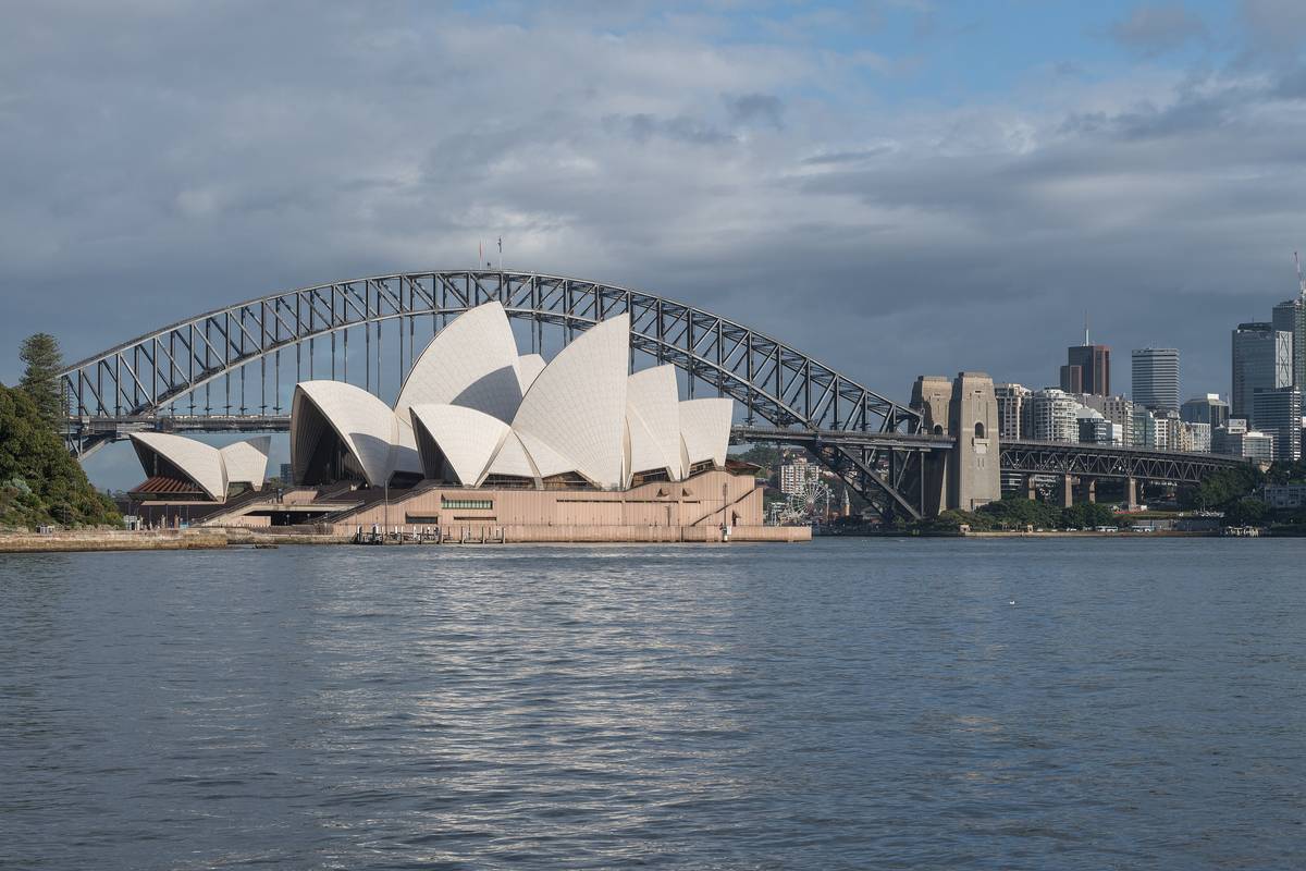 Operahuset i Sydney med sina vita segelformade tak vid vattnet, hamnbron i bakgrunden och skyskrapor under en molnig himmel