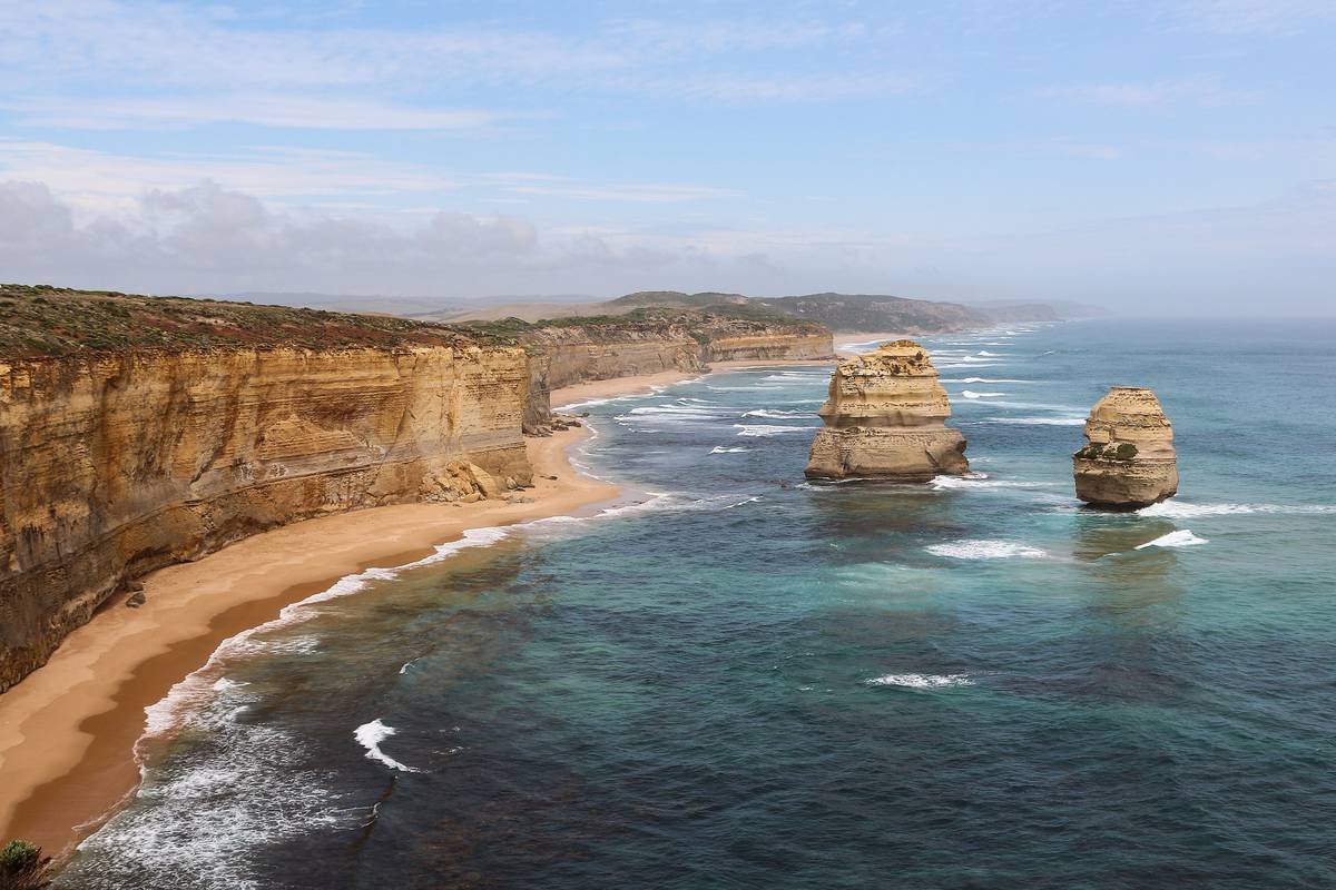 Gula kalkstensklippor och en sandstrand längs kusten, med två höga stenpelare som står ute i det blågröna havet vid Twelve Apostles
