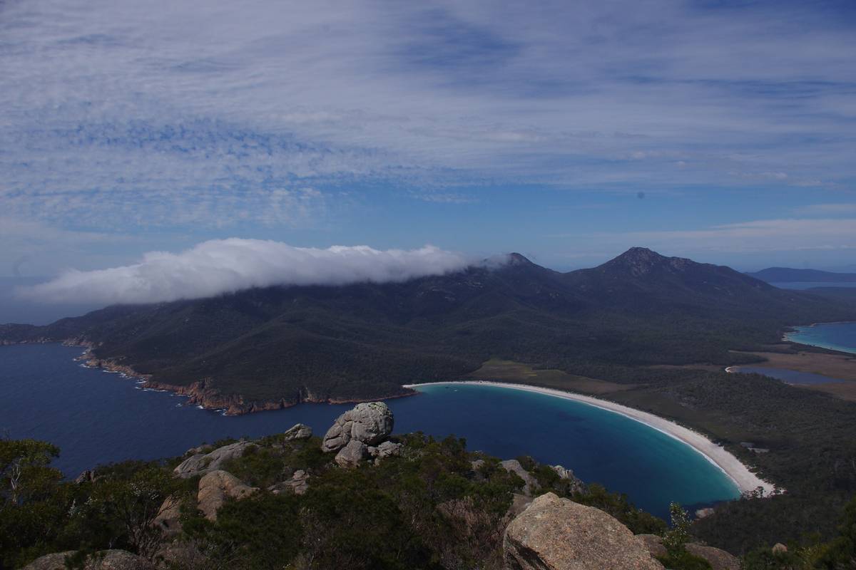 Den halvmåneformade Wineglass Bay med en vit sandstrand och djupblått vatten, omgiven av skogklädda berg under en molnig himmel