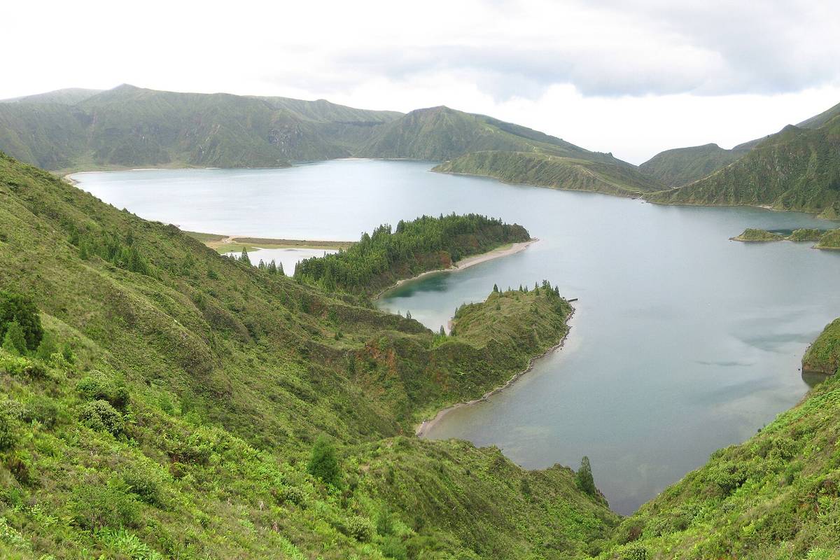 Kratersjön Lagoa do Fogo på São Miguel med en udde och en ljus sandstrand, omgiven av branta gröna sluttningar under en molnig himmel