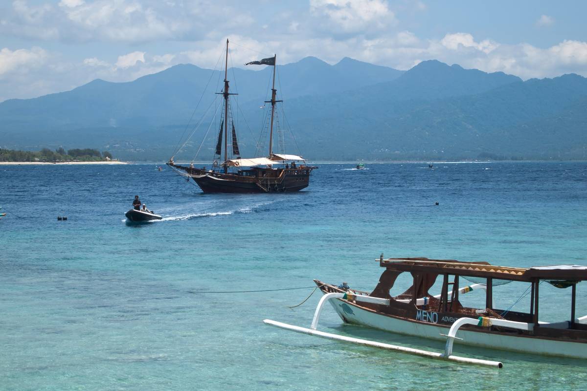 Turkost vatten vid Gili Meno med en utriggarbåt i förgrunden, ett segelfartyg för ankar och Lomboks berg i bakgrunden