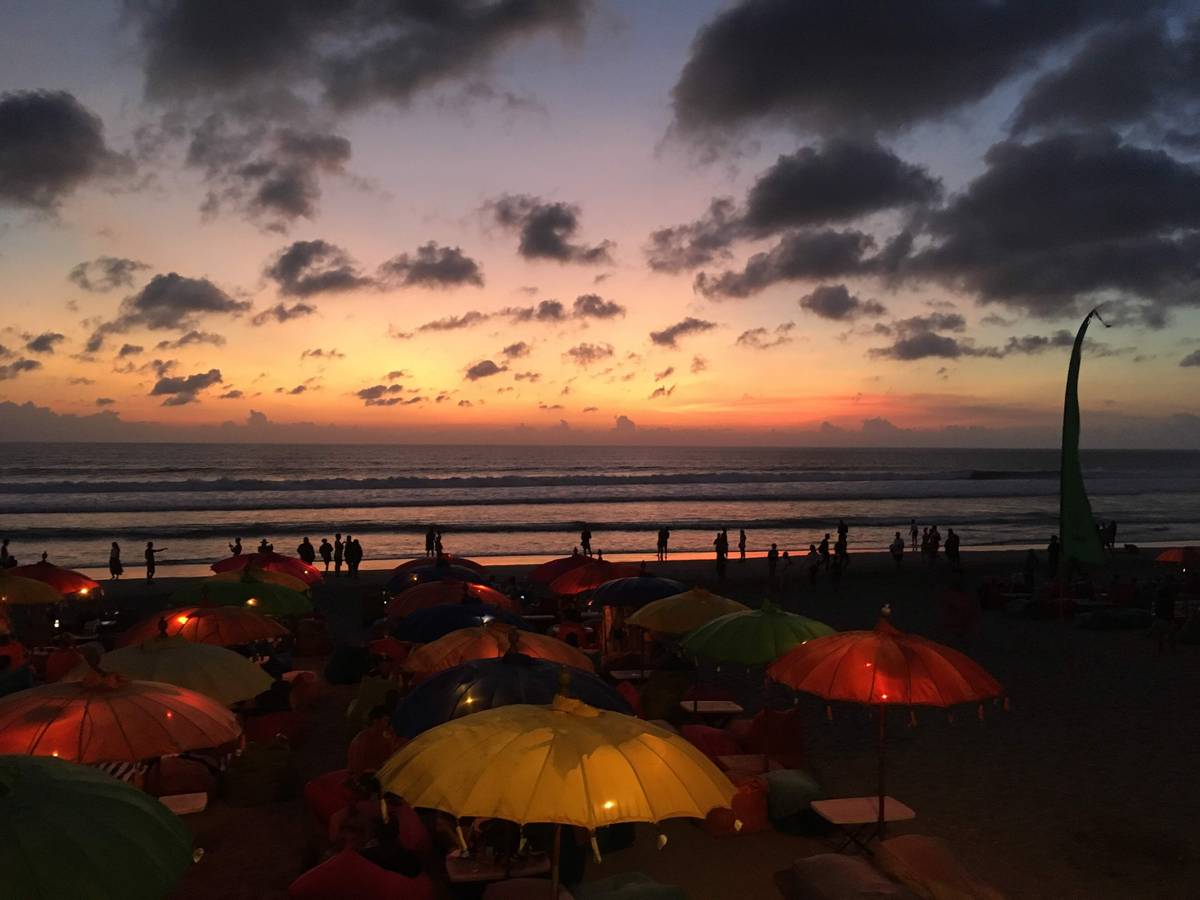 Färgglada parasoller och sittsäckar på stranden i Seminyak i skymningen, med människor i vattenbrynet under en rosa och blå himmel