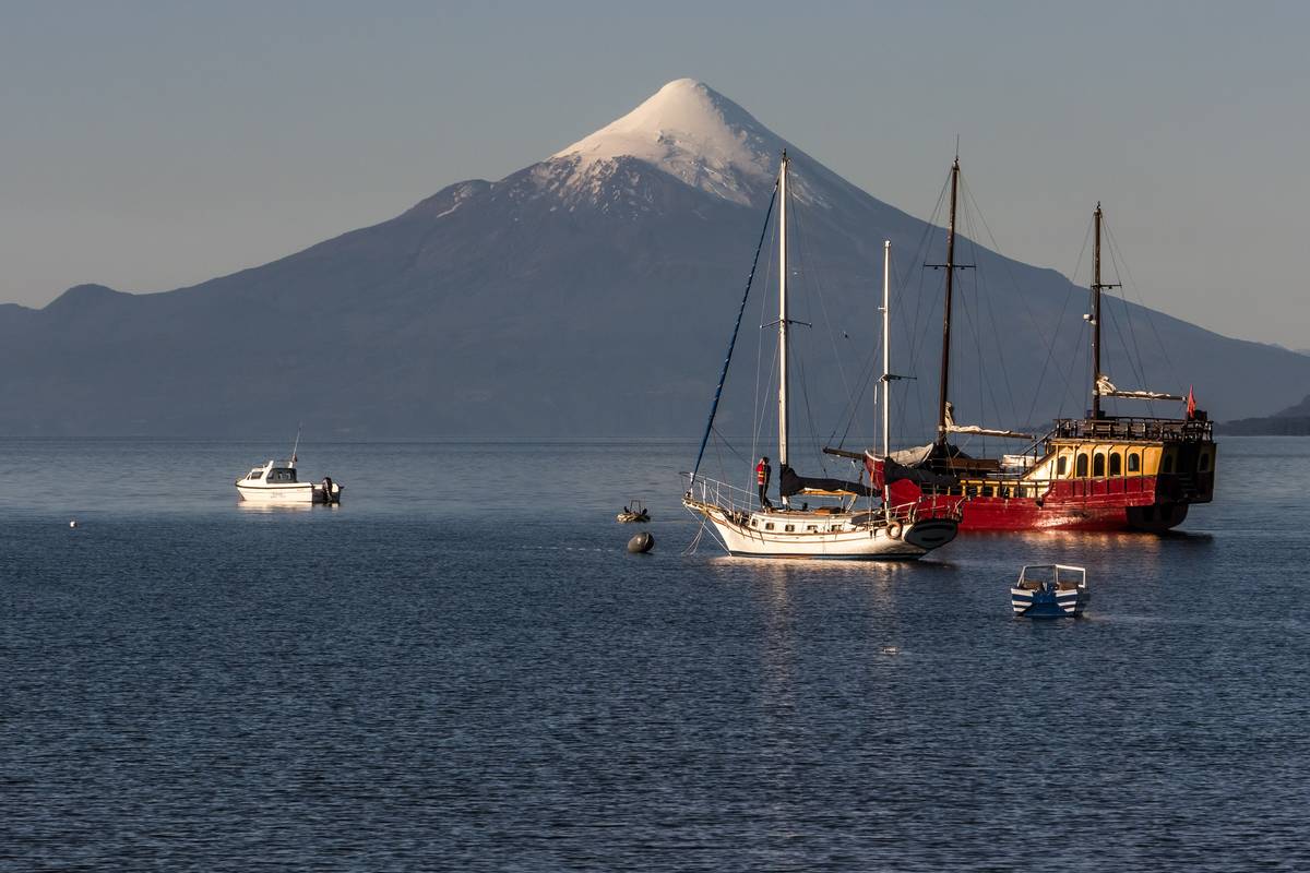Den snötäckta, koniska vulkanen Osorno bakom sjön Llanquihue, där segelbåtar och ett rött skepp ligger för ankar i kvällsljus