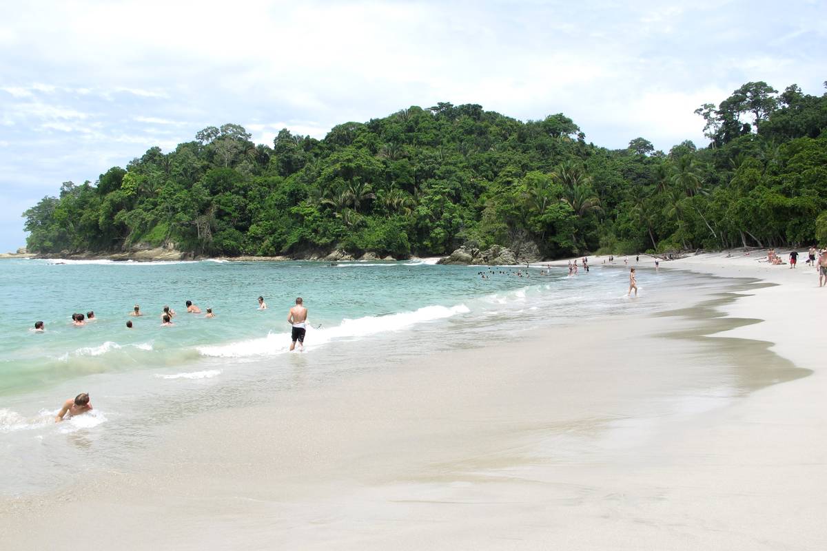 Vit sandstrand med turkosa vågor och badande människor i Manuel Antonio nationalpark, med en skogklädd udde i bakgrunden