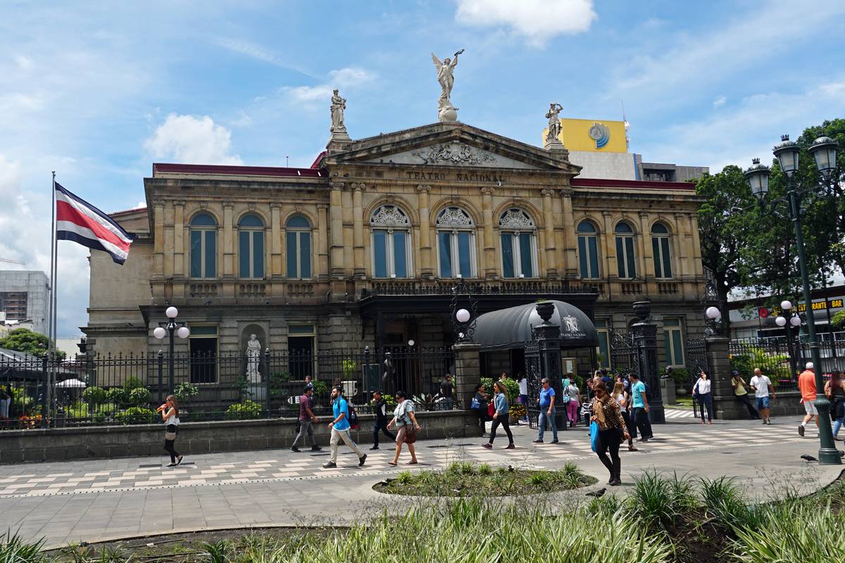 Den gula stenfasaden på Teatro Nacional i San José med statyer på taket, Costa Ricas flagga och folk som går över torget framför