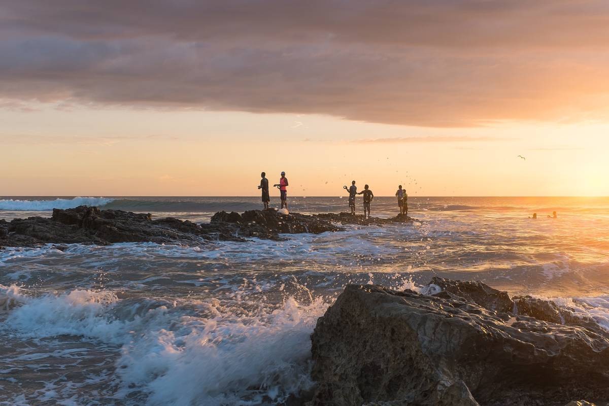Fiskare står på klipporna vid Playa Santa Teresa i solnedgången, med vågor som bryter mot stenarna och en gyllene himmel