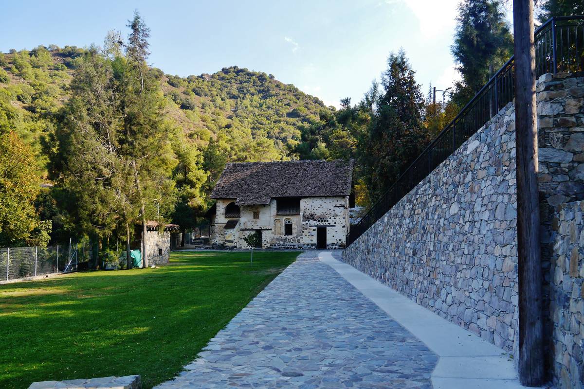 Den lilla stenkyrkan Agios Nikolaos tis Stegis med brant tak, omgiven av grön gräsmatta och skogklädda berg, med en stenmur och en gångväg i förgrunden