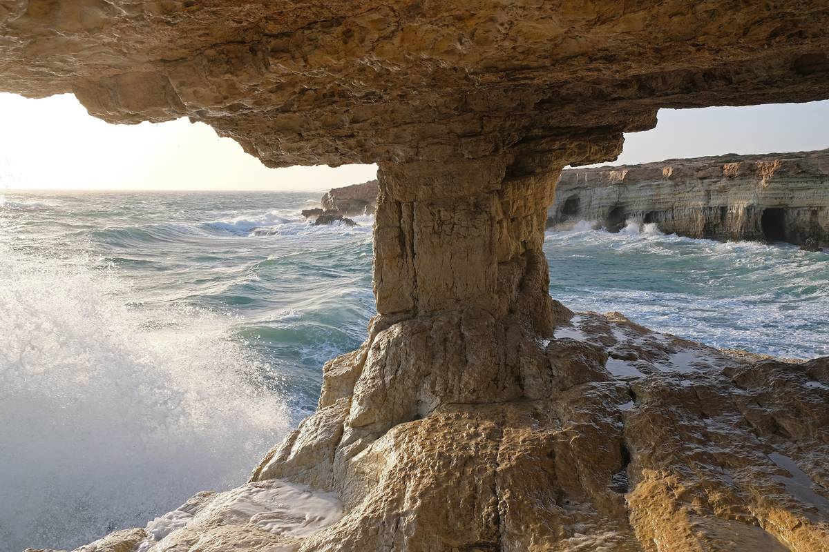 Ett naturligt valv av gulbrun kalksten vid havsgrottorna på Cape Greco, där vågor slår upp mot klipporna och kustlinjen syns i bakgrunden