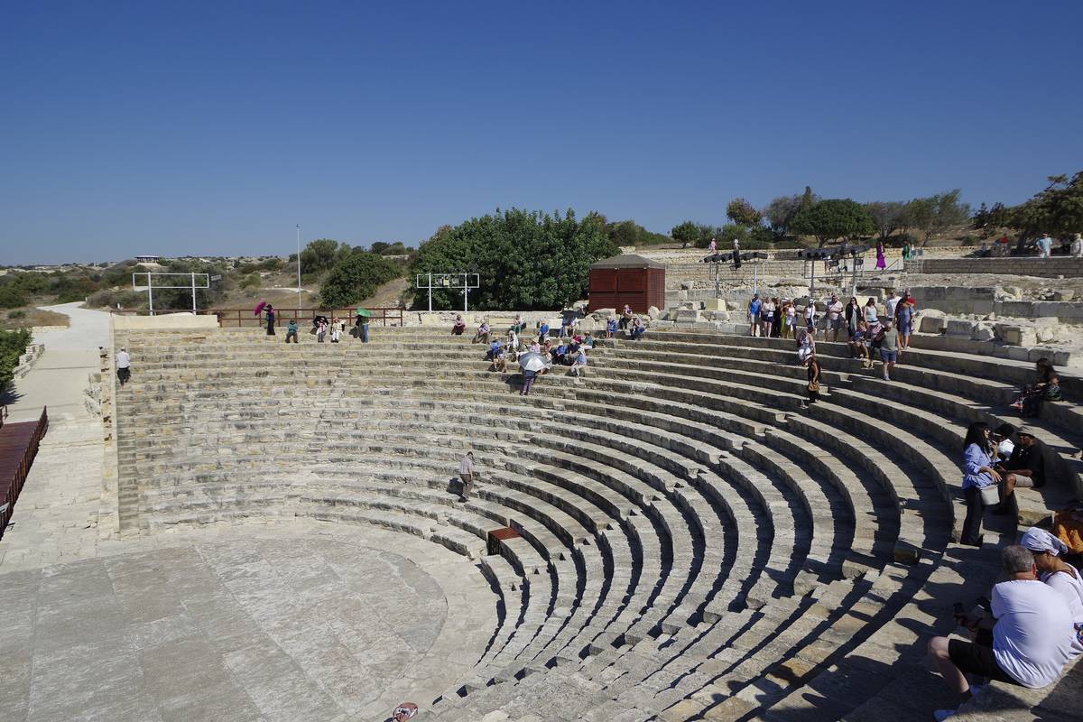 Den antika halvcirkelformade teatern i Kourion med stensittplatser i sluttningen, där besökare sitter och går, under en klarblå himmel