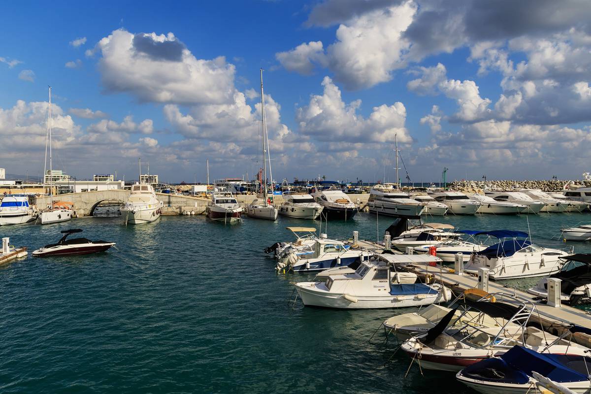 Marinan i Limassol med vita motorbåtar och segelbåtar förtöjda vid bryggor i mörkblått vatten, under en blå himmel med stora vita moln