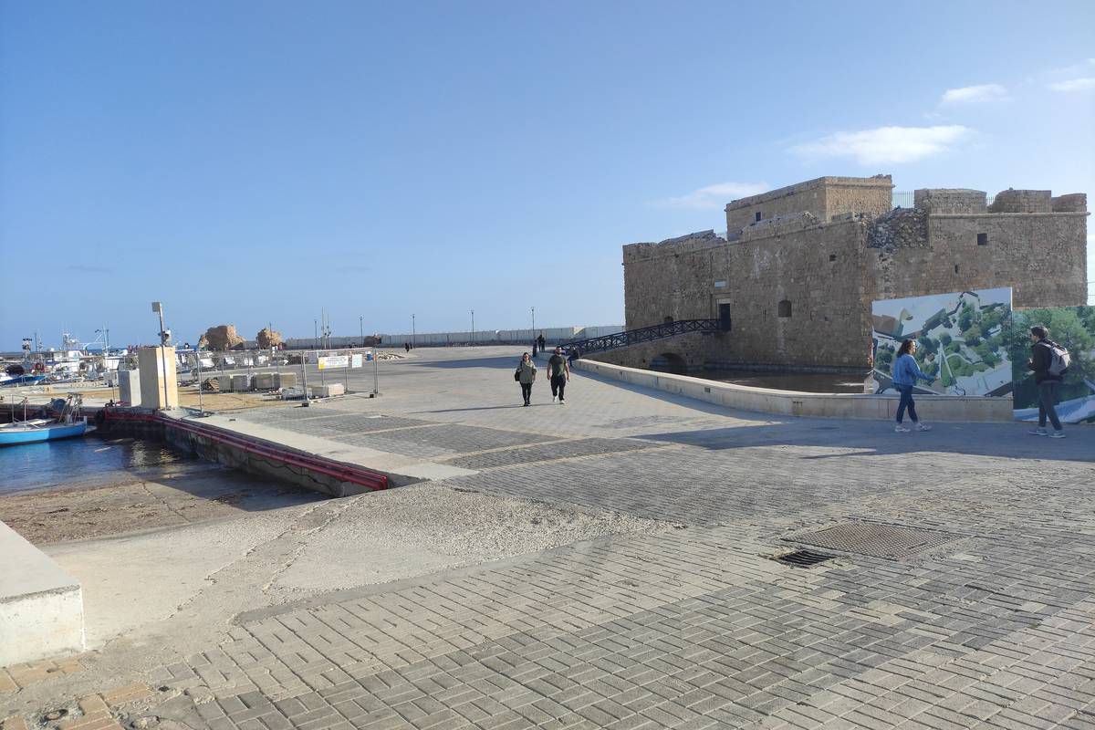 Den medeltida borgen i Pafos, en fyrkantig stenfästning längst ut på hamnpiren, med en bred stenkaj och promenerande människor under en klarblå himmel