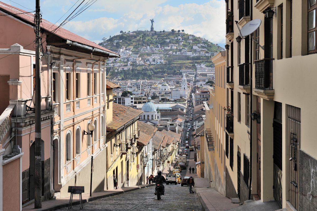 Den smala kullerstensgatan García Moreno i Quitos gamla stad med pastellfärgade kolonialhus och balkonger, och kullen El Panecillo med Jungfrustatyn i bakgrunden