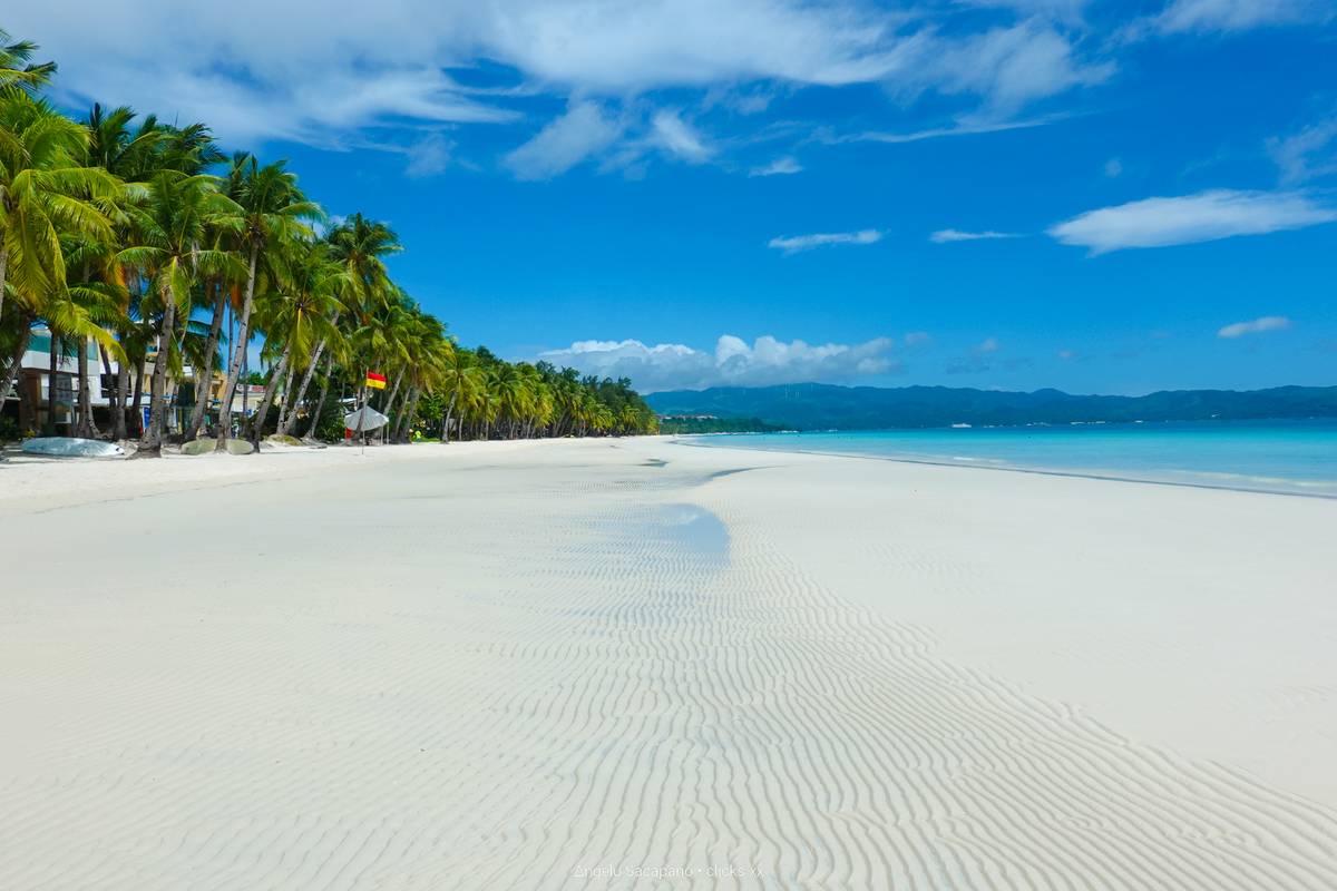 Tom, kritvit sandstrand på White Beach på Boracay med krusningar i sanden, en rad palmer och turkost vatten under blå himmel