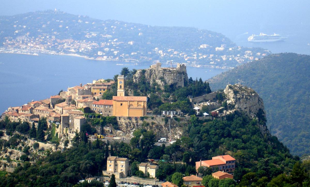Den medeltida byn Èze på en klippa ovanför Medelhavet, med stenhus och ett gult kyrktorn, gröna sluttningar och udden Cap Ferrat i bakgrunden