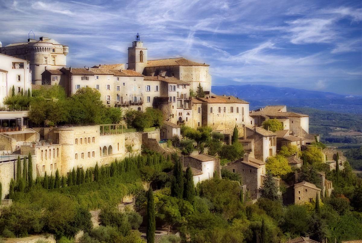 Byn Gordes i Luberon med ljusa stenhus och ett slott på toppen av en kulle, cypresser och gröna träd nedanför och fjäderlika moln på himlen