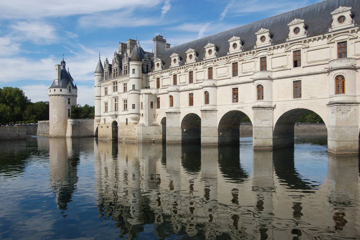 Slottet Chenonceau i vit sten med valv över floden Cher, spegelblankt vatten som speglar fasaden och ett runt torn till vänster under blå himmel