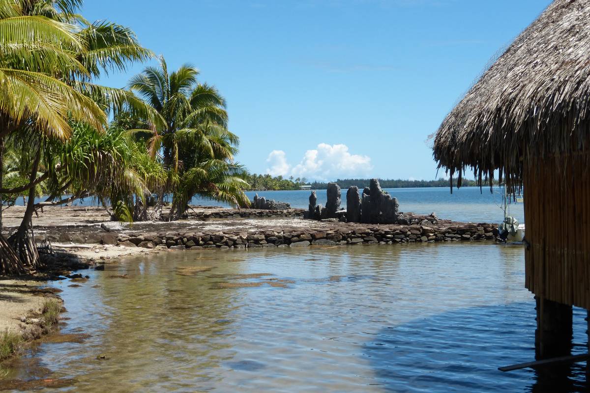 Stenmurar från en marae vid Maeva på Huahine, vid grunt klart vatten med palmer och ett halmtak i förgrunden