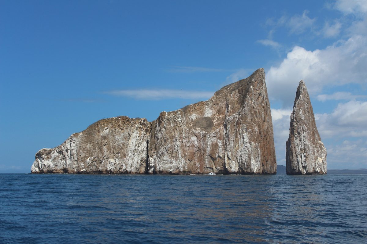 Den branta klippan Kicker Rock reser sig ur det djupblå havet i två delar med en smal kanal emellan, under en blå himmel med vita moln