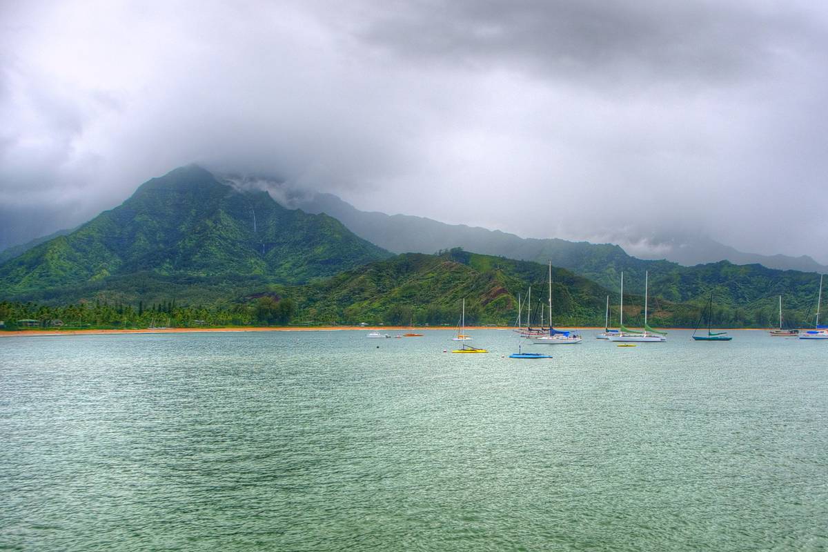 Segelbåtar för ankar i den lugna, grönblå Hanalei Bay på Kauaʻi, med frodiga gröna berg och låga moln bakom