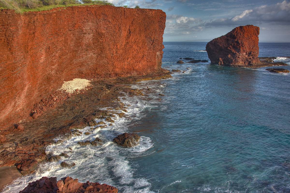 Den rödbruna klippön Puʻu Pehe som reser sig ur det blå havet bredvid en hög röd lavaklippa på Lānaʻis sydkust, med vita vågor mot stenarna
