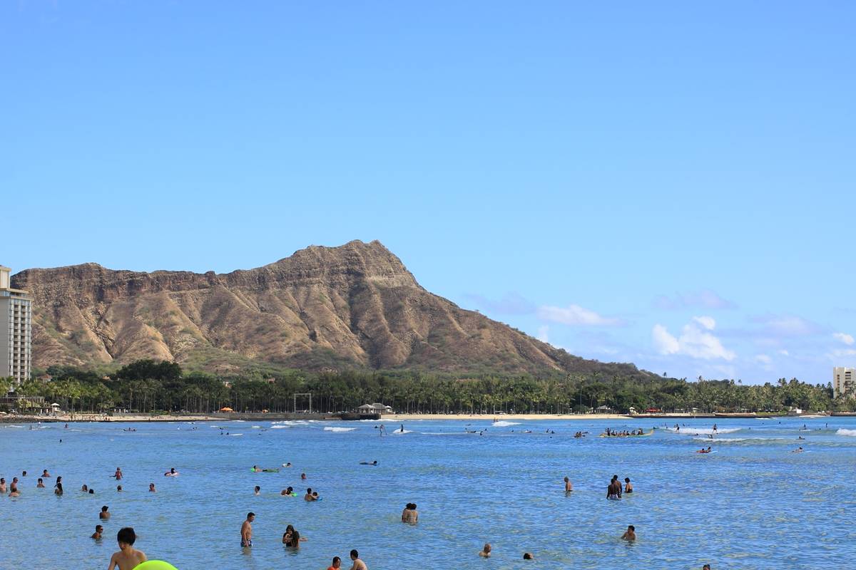 Badande i det lugna, blå vattnet vid Waikīkī Beach, med den bruna kraterkanten Diamond Head och gröna palmer bakom under en klarblå himmel
