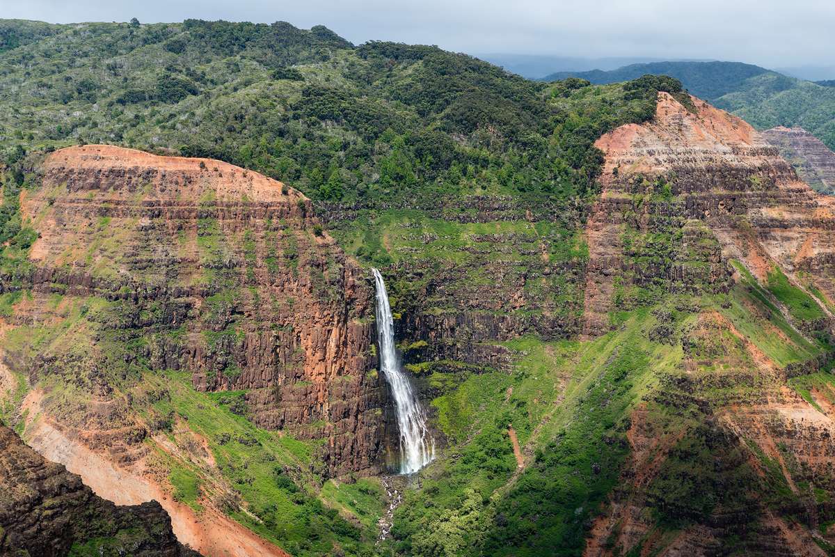 Vattenfallet Waipoʻo som störtar ner mellan röda och gröna klippväggar i Waimea Canyon, med skogklädda bergskammar runt omkring