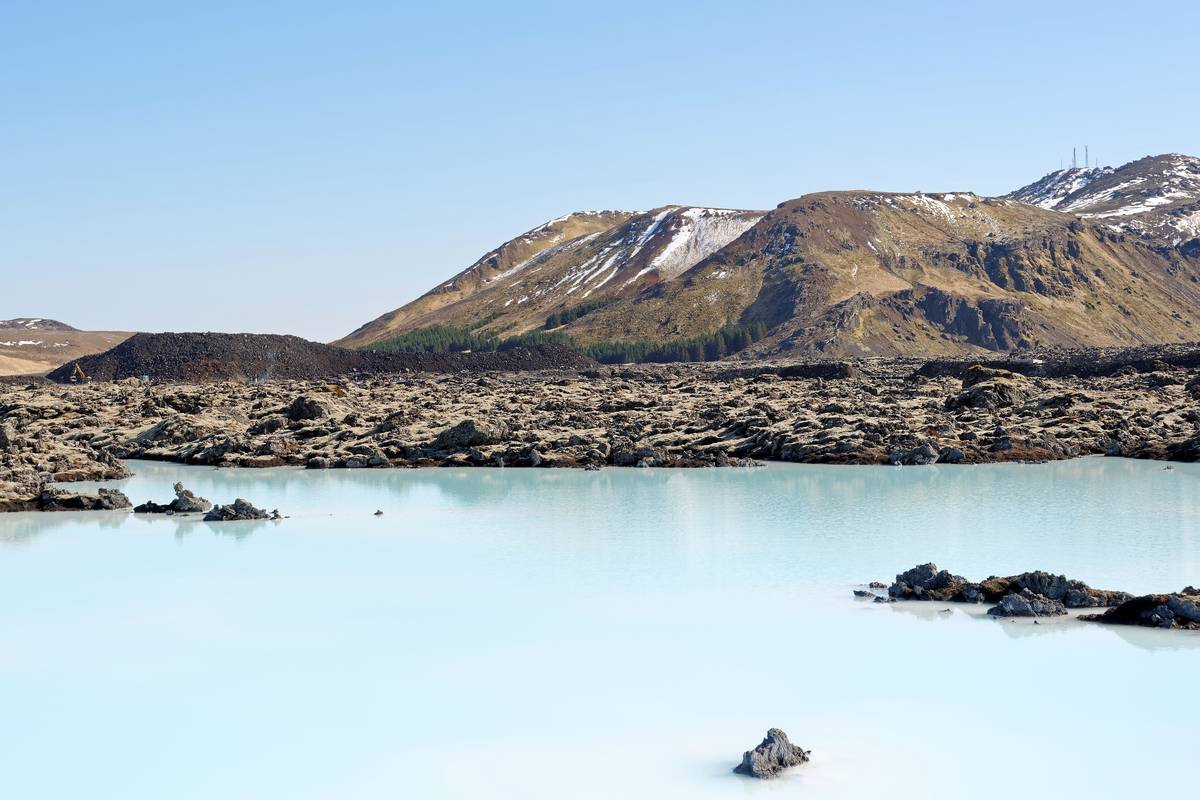 Det mjölkblå vattnet i Blå lagunen mellan svarta lavastenar, med berget Þorbjörn bakom under en klarblå himmel