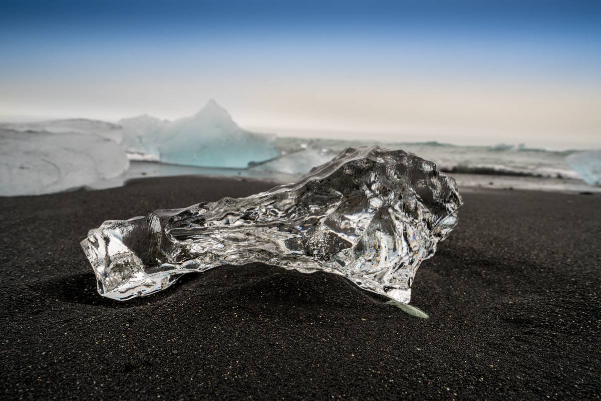 Ett genomskinligt isblock som glittrar på den svarta sanden på Diamond Beach, med havet i bakgrunden