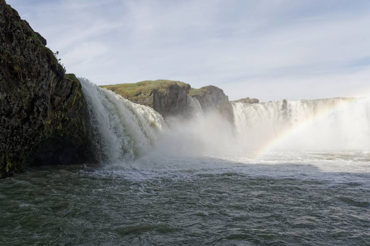 Det breda, hästskoformade vattenfallet Goðafoss med en regnbåge i vattendimman och mörka klippor runt omkring