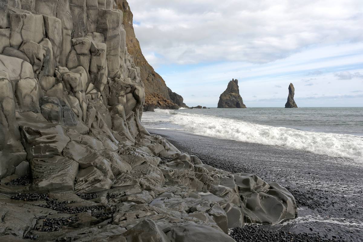 Mörka basaltpelare på stranden Reynisfjara, med vita vågor mot den svarta sanden och klipporna Reynisdrangar ute i havet