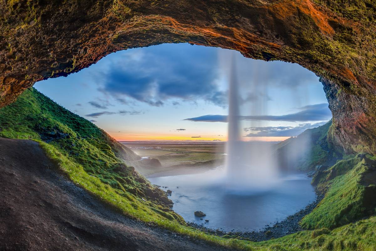 Seljalandsfoss sett inifrån grottan bakom vattenfallet, där vattnet faller ner framför gröna slätter och en kvällshimmel i orange och blått