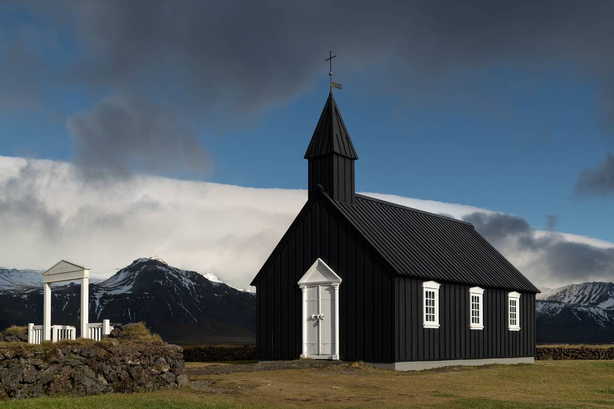 Den svarta träkyrkan Búðakirkja med vita fönster, en stenmur framför och berg under mörka moln bakom