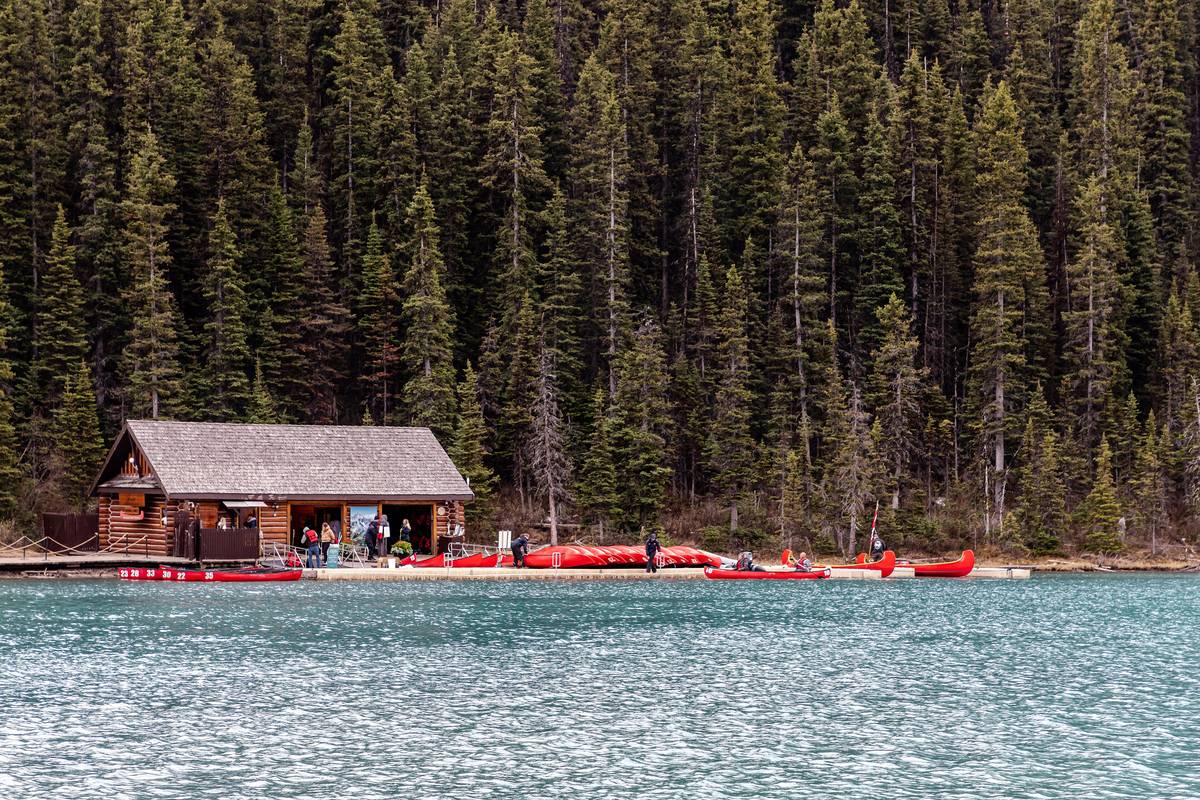 Ett brunt timrat båthus med röda kanoter på bryggan vid den grönblå Lake Louise, framför en tät granskog