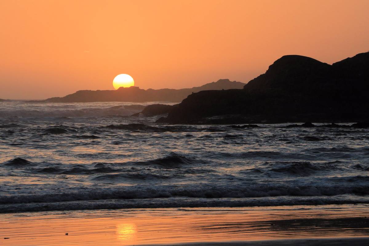 Solen går ner bakom en mörk, skogklädd udde vid Cox Bay nära Tofino, med orange himmel och vågor som rullar in över stranden