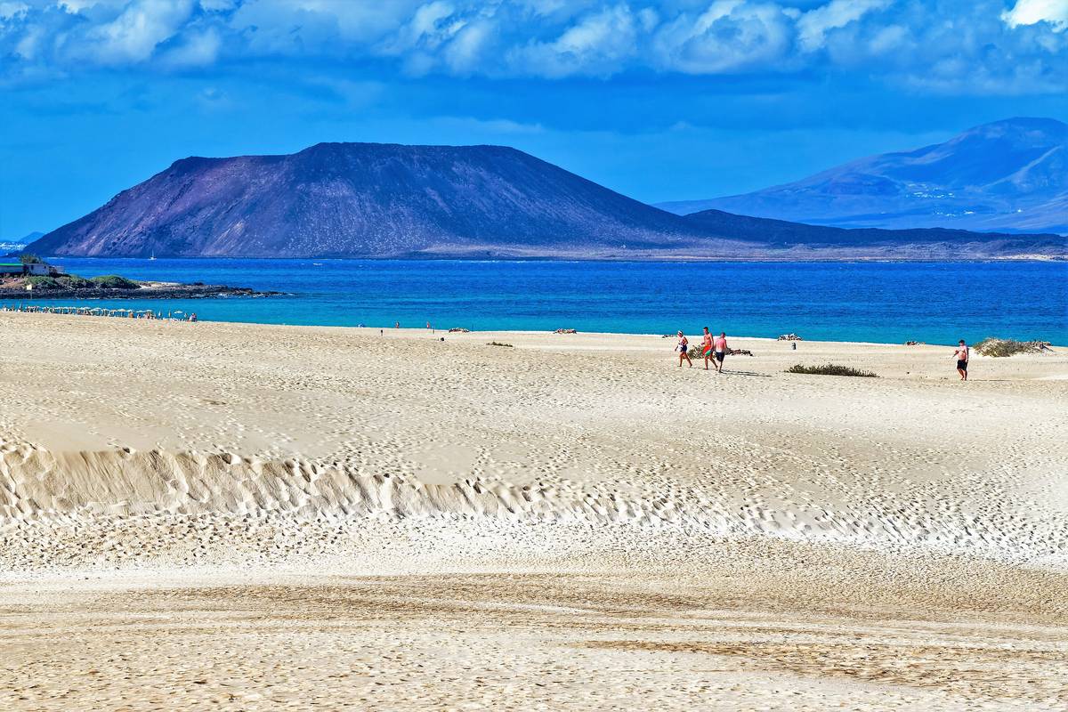 Vita sanddyner i Corralejo på Fuerteventura som möter ett turkost hav, med några strandvandrare och den bruna vulkanön Lobos bakom