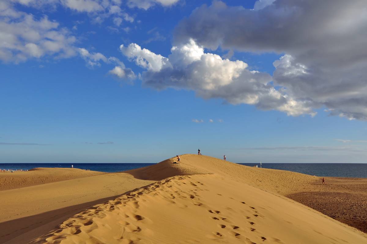 Gyllene sanddyner i Maspalomas på Gran Canaria med fotspår i sanden, några små människor på krönet och havet och vita moln bakom