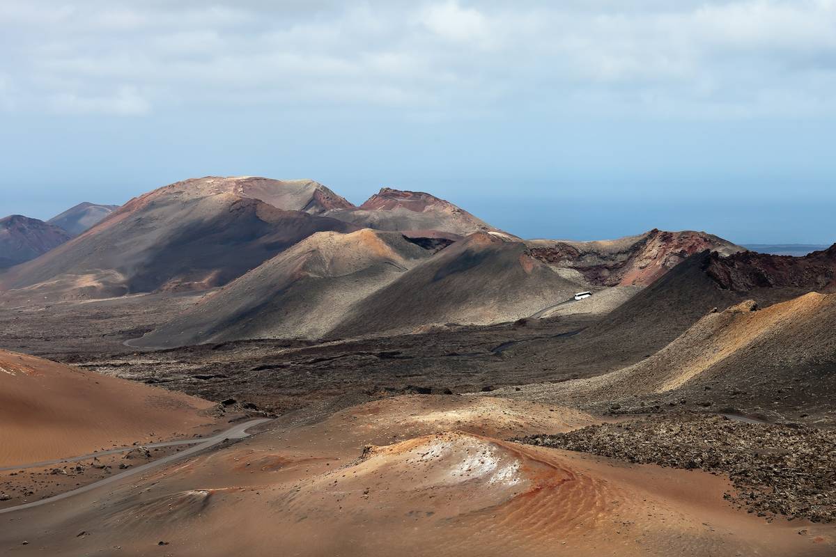 Vulkankäglor i rött, brunt och ockra i Timanfaya nationalpark på Lanzarote, med en slingrande väg genom lavafälten och havet vid horisonten