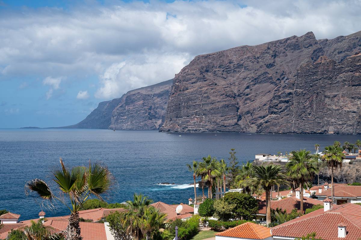 De mörka, lodräta klipporna vid Los Gigantes på Teneriffa som stupar ner i ett djupblått hav, med palmer och hus med röda tegeltak i förgrunden