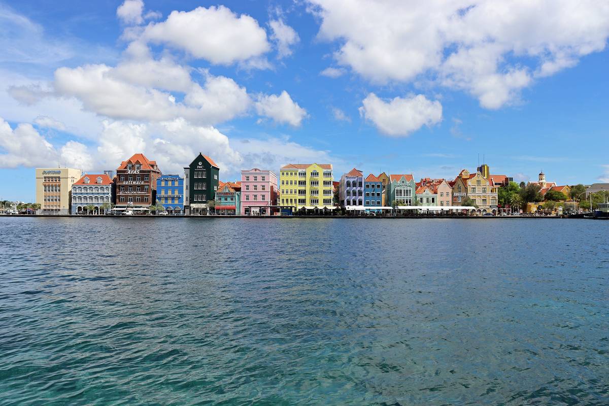 Rad av pastellfärgade kolonialhus i holländsk stil längs vattnet vid Handelskade i Willemstad under blå himmel med vita moln