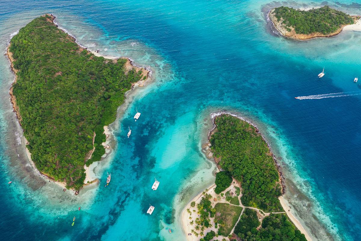 Flygbild över gröna, obebodda öar med vita stränder och ankrade segelbåtar i turkost, grunt vatten i Tobago Cays