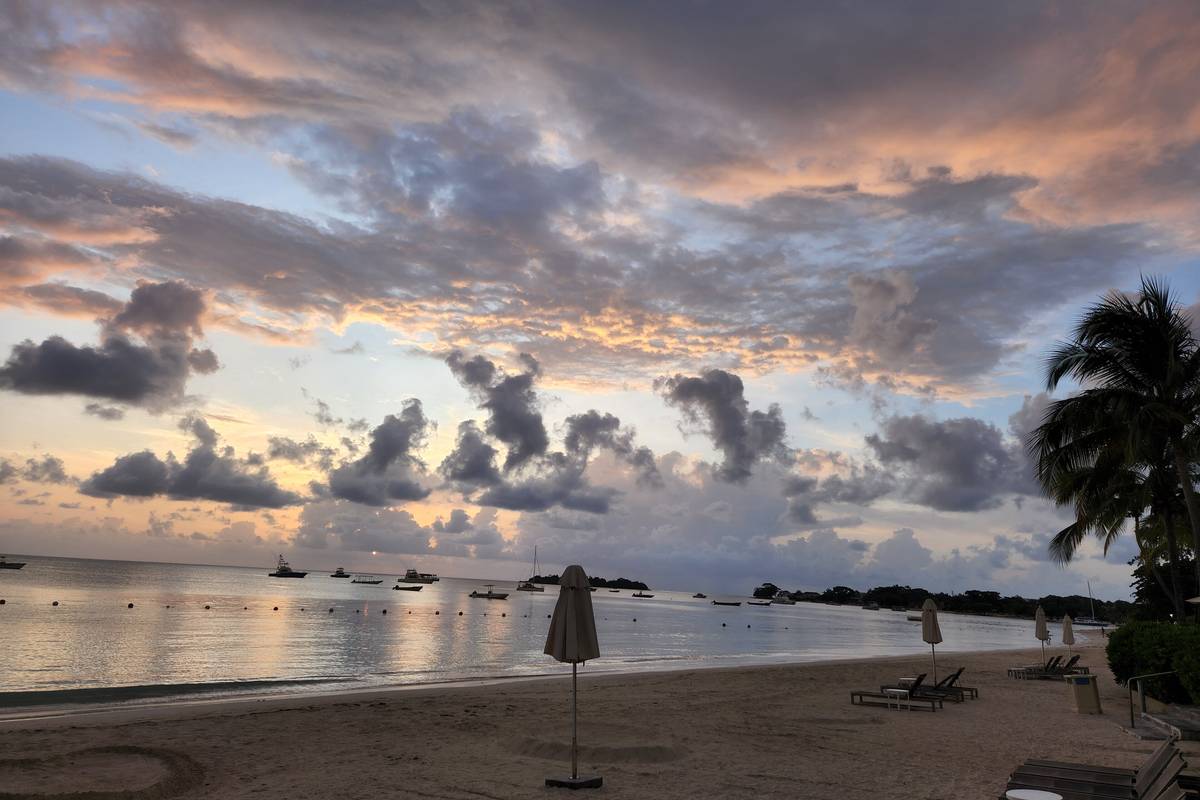 Seven Mile Beach i Negril i skymningen, med ett parasoll på sanden, en palm och båtar på det stilla vattnet under rosa moln