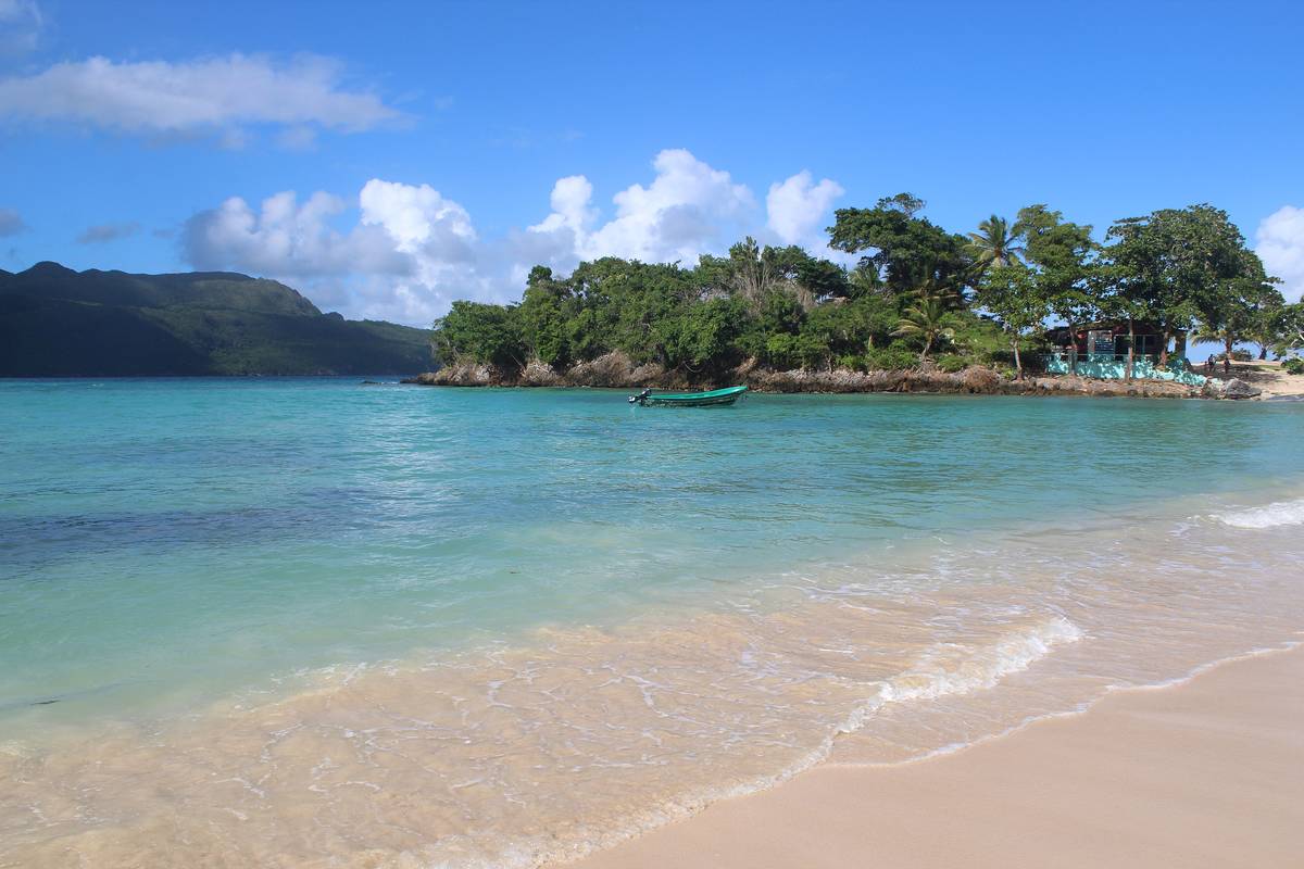 Ljus sandstrand och turkost, klart vatten vid Playa Rincón, med en grön udde, en liten båt och skogsklädda berg i bakgrunden