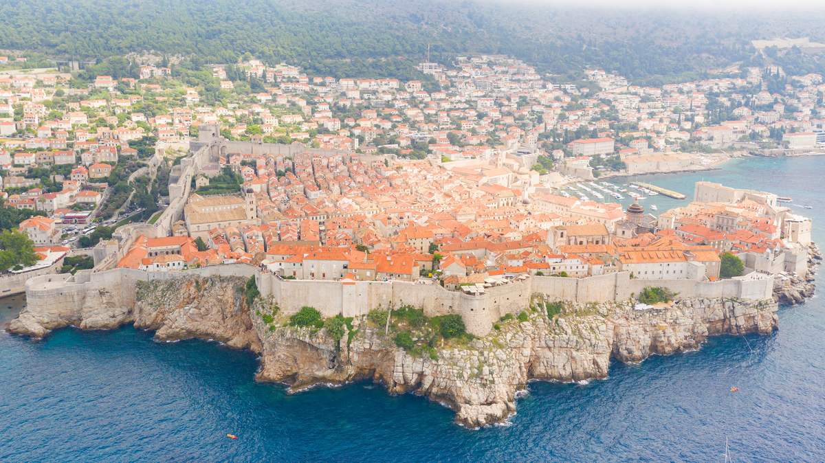 Flygbild över Dubrovniks gamla stan med röda tegeltak innanför den ljusa stadsmuren, som följer den branta klippkusten ner mot havet