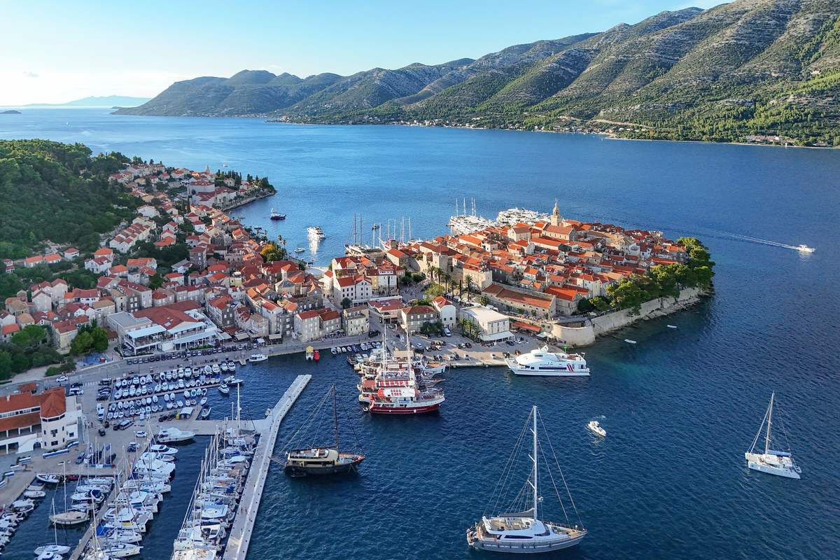 Flygbild över Korčulas gamla stan på en udde med röda tak, en hamn med segelbåtar och Pelješacs berg på andra sidan sundet