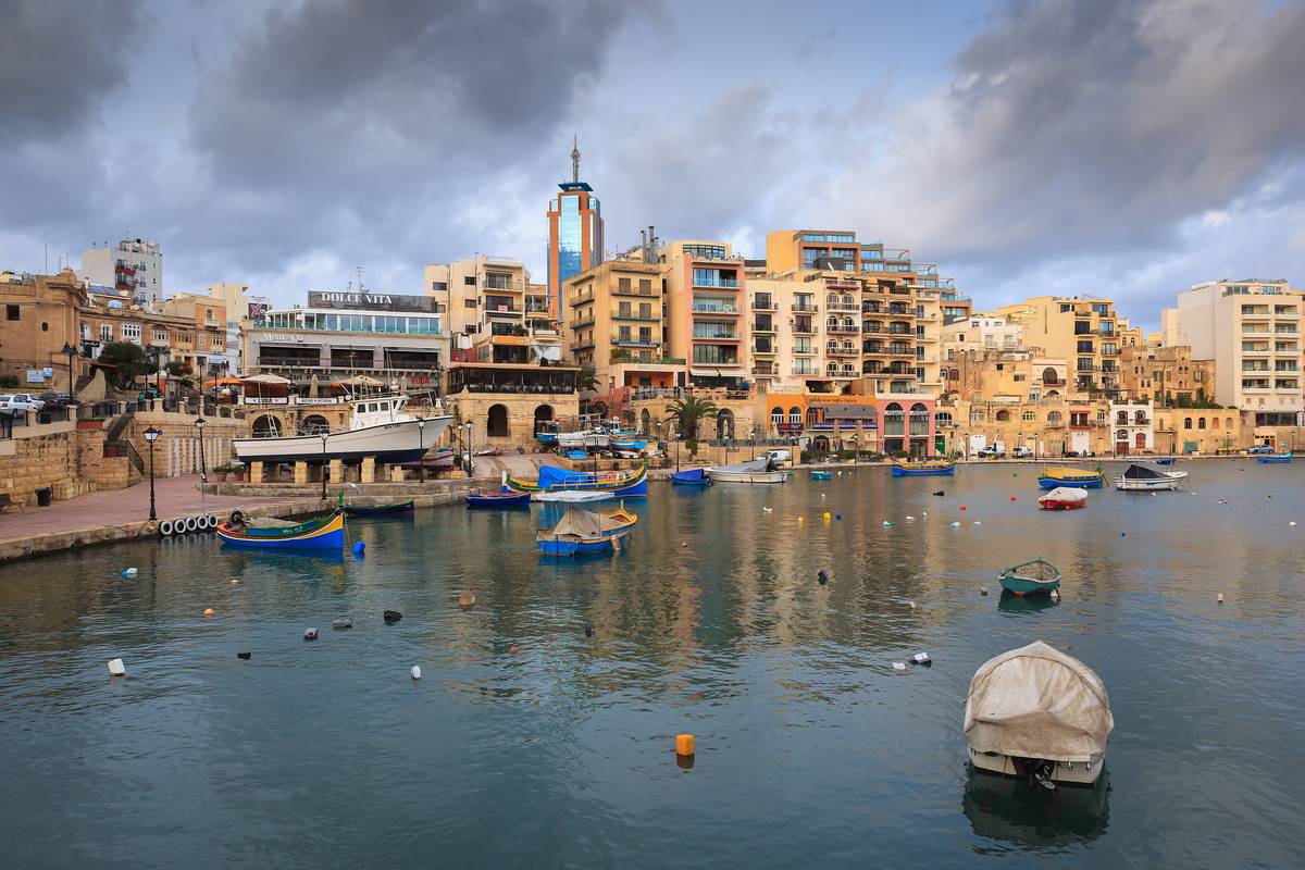 Spinola Bay i St Julian's med små fiskebåtar på det stilla vattnet och restauranger och hus runt viken under en dramatisk himmel