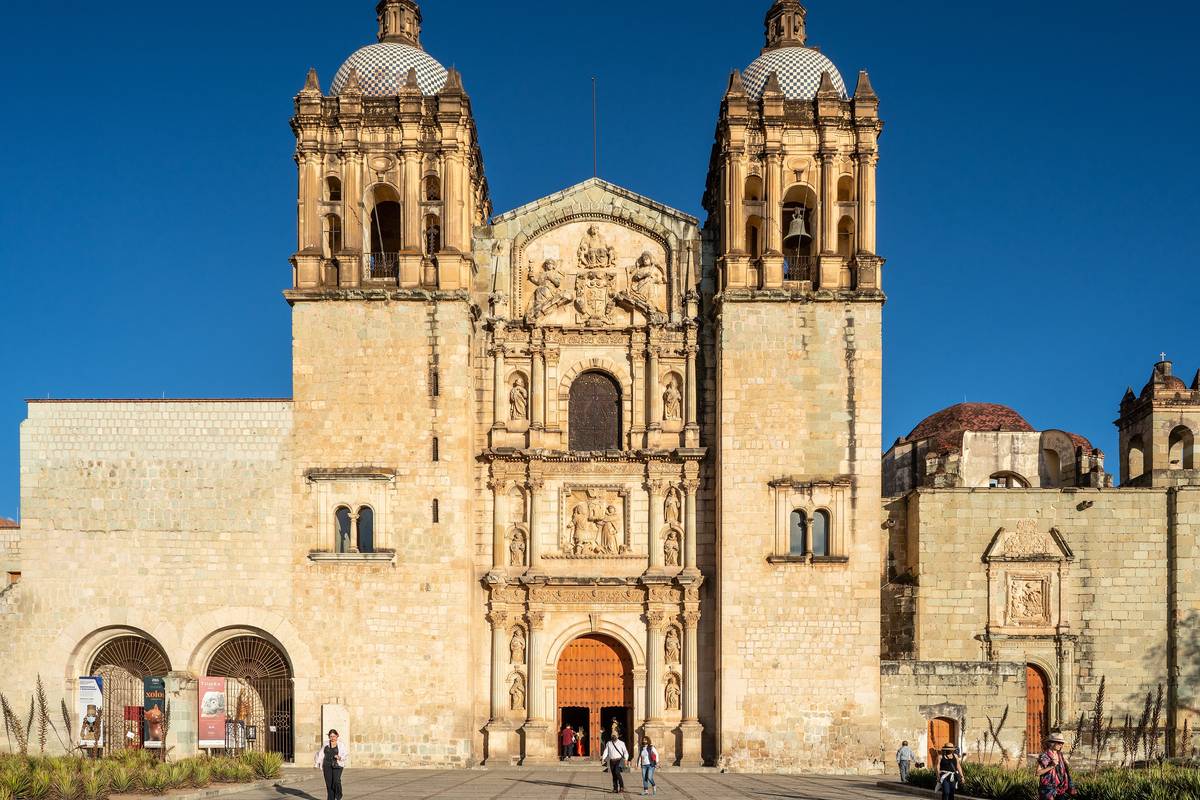 Kyrkan Santo Domingo de Guzmán i ljus sten med två klocktorn med kupoler och en rikt utsmyckad fasad under klarblå himmel i Oaxaca